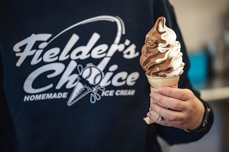 Person holding a chocolate and vanilla swirl ice cream cone in front of Fielder's Choice branded sweatshirt.